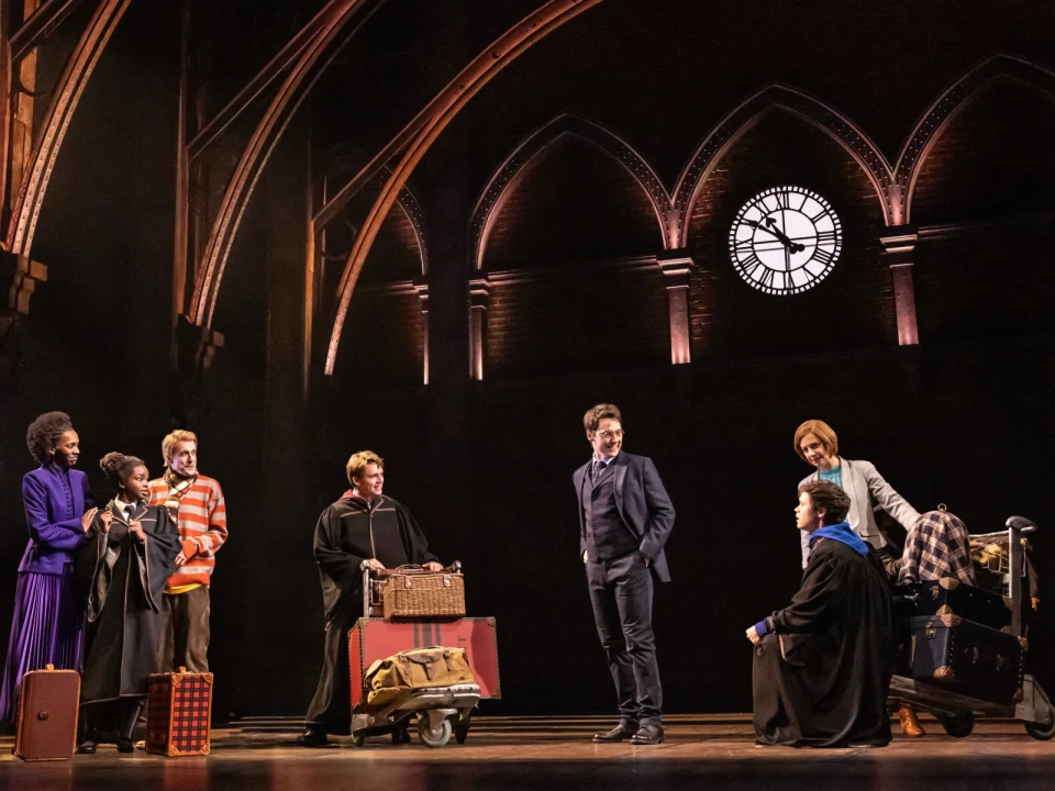 A group of actors performs a scene on stage with a large clock in the background. They are dressed in costumes and holding luggage, suggesting a travel or departure theme.