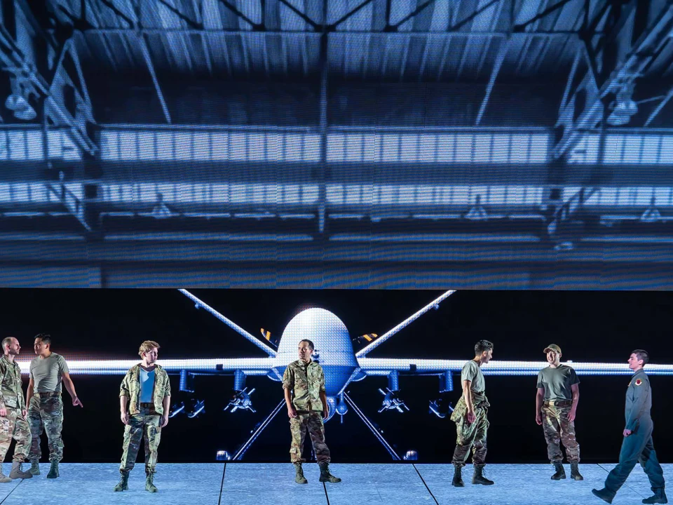 A group of people in military uniforms stand on stage with a large screen displaying an aircraft hangar and a drone in the background.