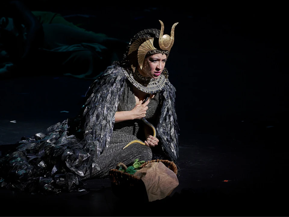 A performer in an elaborate costume with a headdress and feathered cloak kneels on stage holding a snake prop, with a basket in front of them.