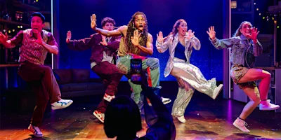 Five performers are mid-dance on a brightly lit stage, each with an expressive pose, as someone in the foreground takes a photo or video with a phone.