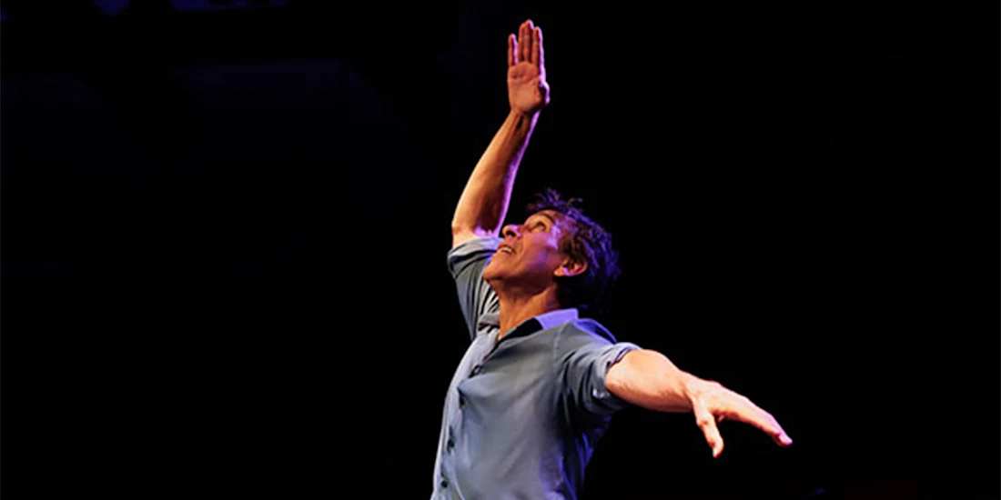 A person in a blue shirt extends one arm sideways and raises the other arm upward against a dark background.