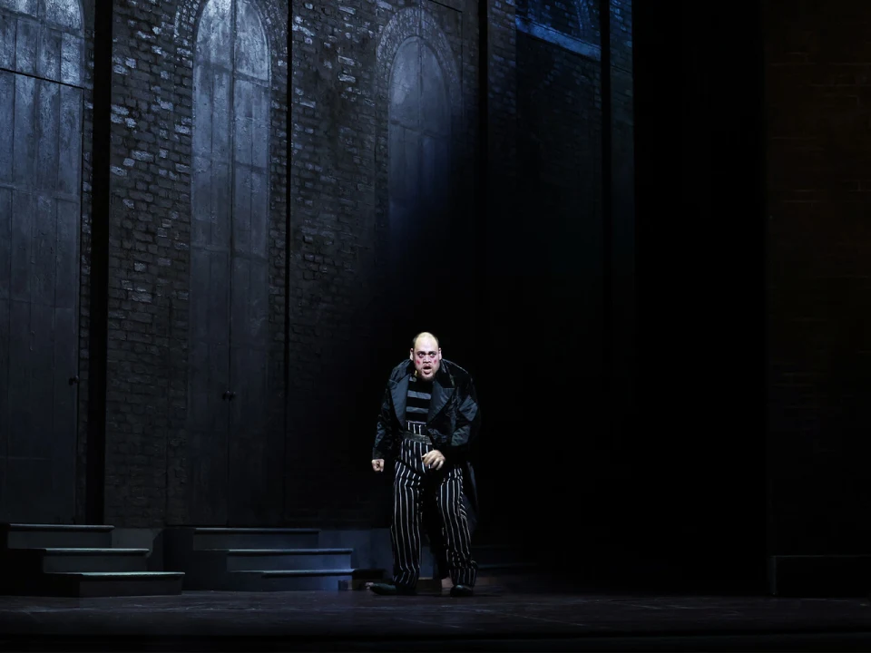 A person dressed in dark, striped clothing stands on a dimly lit stage with brick walls and arched doorways in the background.