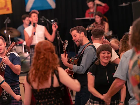 A group of musicians and performers play guitars and sing together on stage, surrounded by audience members in Scottish dress.
