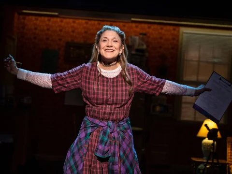 A woman with arms outstretched stands on stage, smiling broadly. She is dressed in a plaid outfit and holds a clipboard in one hand. The background features a warmly lit, homely interior setting.