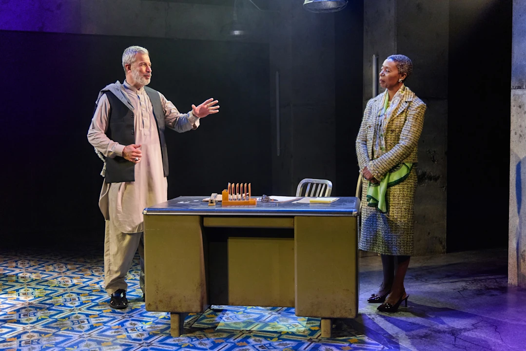 A man gestures while speaking to a woman who stands beside a desk in a dimly lit room with patterned floor tiles.