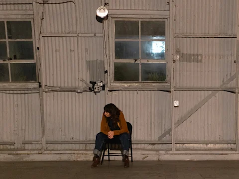 A person sits alone on a chair in front of a corrugated metal wall with two windows and visible utility fixtures.