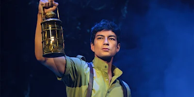 A person in a green shirt holds up a brass lantern, looking ahead with a serious expression against a dark, misty background.