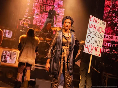 Production photo of Rolling Thunder in New York showing a woman on stage holds a protest sign reading "END THE WAR NOW. BRING OUR SONS HOME!" with musicians and performers in the background.