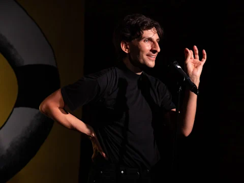 A person in a black shirt stands on stage speaking into a microphone, with one hand on their hip and the other gesturing, against a dark background with a yellow and white design.