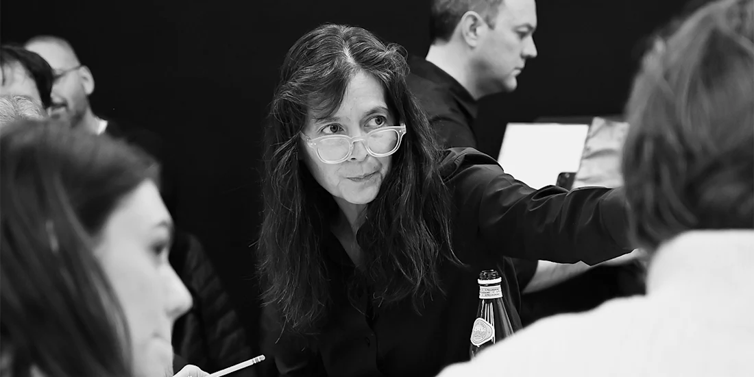 A woman with glasses gestures while speaking to others in a group setting; several people are visible around her in a black-and-white scene.