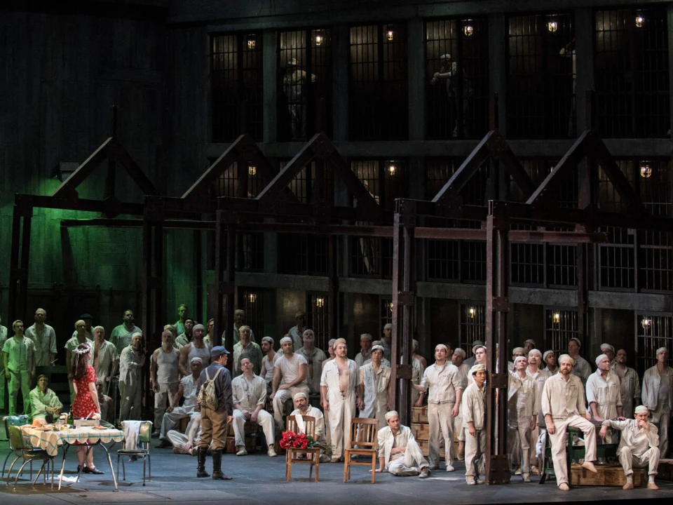 A large group of people in white clothing stand in an industrial setting with metal bars and dark walls. The scene is dimly lit with a woman in a red dress standing to the left.