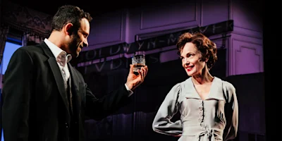 A man in a suit holds up a glass of whiskey while facing a woman in a light-colored dress, both standing on a dimly lit stage.