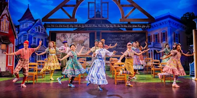 Actors in colorful costumes perform a lively dance on stage set as a classroom, with a chalkboard and wooden chairs, framed by painted historic houses and blue sky backdrop.