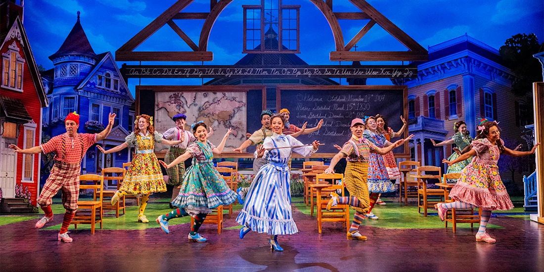Actors in colorful costumes perform a lively dance on stage set as a classroom, with a chalkboard and wooden chairs, framed by painted historic houses and blue sky backdrop.