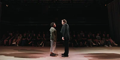 Two people stand facing each other on a stage with an audience seated in the background, under dim lighting.