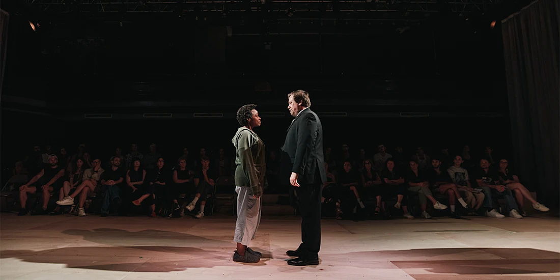 Two people stand facing each other on a stage with an audience seated in the background, under dim lighting.