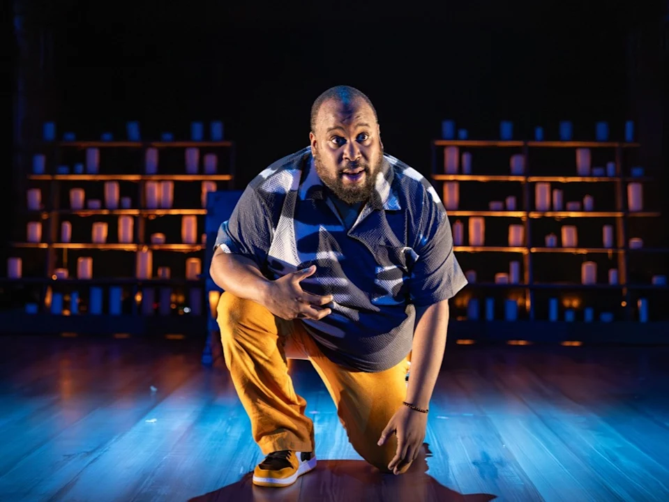 A man kneels on a stage under dramatic lighting, with bookshelves and illuminated candles in the background.