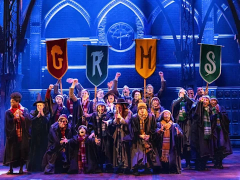 Production photo of Harry Potter and the Cursed Child in Costa Mesa showing a theater cast dressed as Hogwarts students stands on stage, holding banners for the four houses, with arms raised and excited expressions under blue lighting.