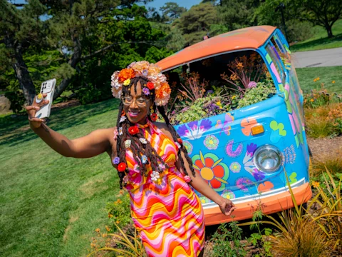 A woman in a colorful flower-print dress and floral headpiece takes a selfie in front of a brightly painted van decorated with flowers in a park.