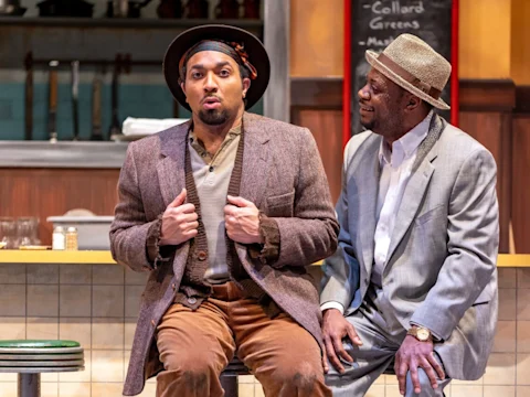 Two men sit on barstools in a diner setting. One wears a brown hat and suit, while the other, with a bandana and brown jacket, gestures as if talking or performing.