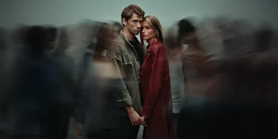 A man and woman stand close together holding hands, facing the camera, surrounded by blurred people in motion.