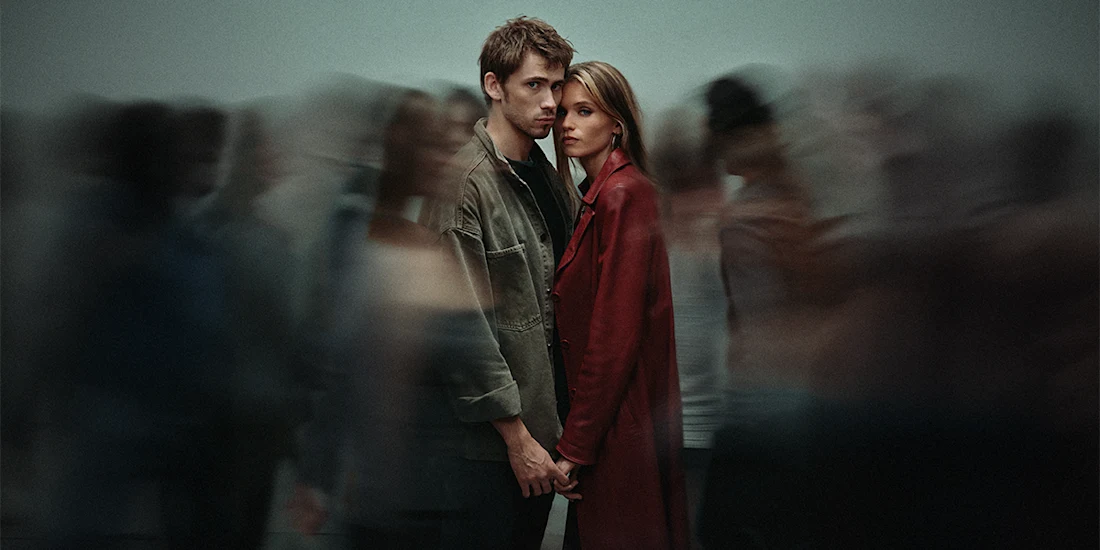 A man and woman stand close together holding hands, facing the camera, surrounded by blurred people in motion.
