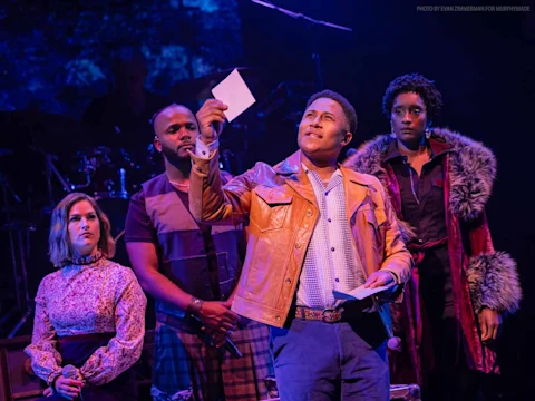 Production photo of Rolling Thunder in New York showing four actors in period costumes stand on stage; one in a tan jacket holds up a piece of paper, appearing to address the audience, with dramatic lighting highlighting the scene.