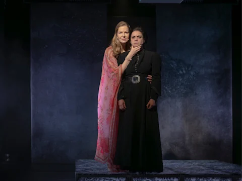 Two women stand on a dimly lit stage, one in a pink floral shawl and the other in a black dress, against a textured dark background.