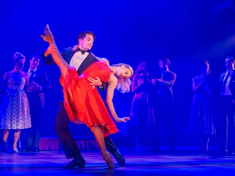 A man in a tuxedo dips a woman in a red dress on stage, with other performers in vintage attire standing in the background.