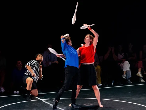 Two jugglers in blue and red shirts perform a routine with clubs on a stage, while a referee watches closely in the background. An audience sits in darkness at the back.