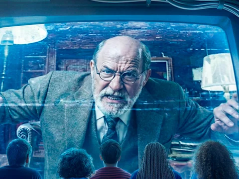 Four people sit facing a large screen displaying an older man with glasses and a serious expression, wearing a suit and leaning forward in an office setting.