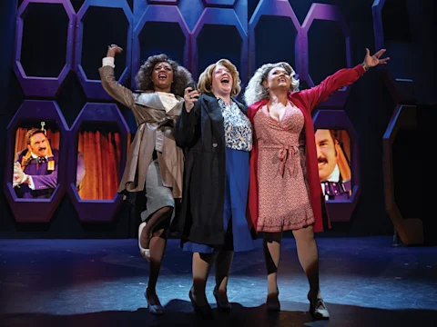 Three women in professional and casual clothing stand together on stage, smiling and singing, with purple hexagonal frames and projected images in the background.