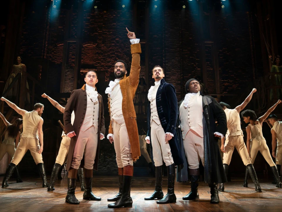 Four men in historical costumes stand together on stage with a group in the background, set in a theatrical performance with dramatic lighting.