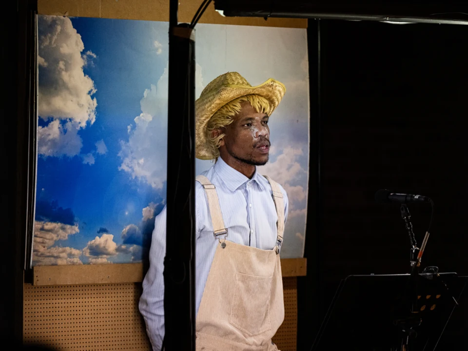A person wearing a straw hat and overalls stands in front of a sky backdrop, speaking into a microphone.