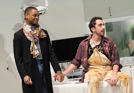 Two people hold hands in a room with medical equipment. One wears a scarf and coat, the other is in a paint-splattered outfit, sitting on a chair.