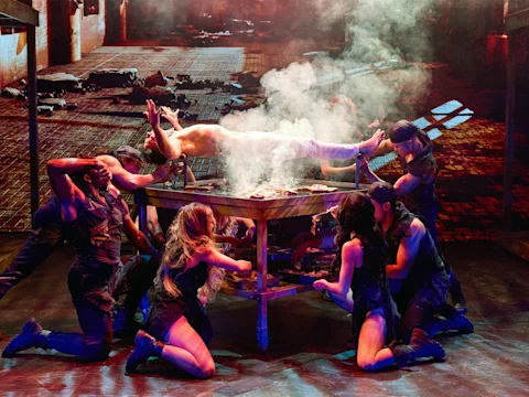 A shirtless person lies on a smoking table while six performers in dark costumes pose around the table on stage, with a dramatic, industrial backdrop.
