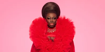 A person with dramatic makeup and a large beehive hairstyle wears a bright red ruffled garment against a pink background.
