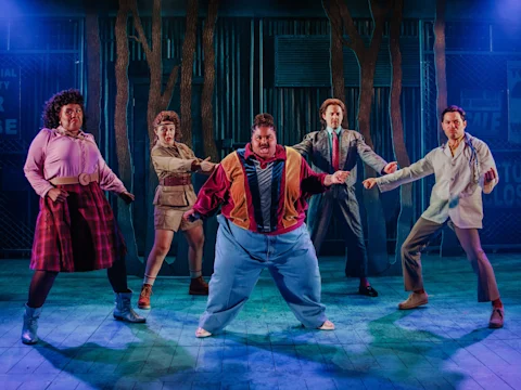 Five actors in colorful, retro costumes stand in powerful poses on stage with dramatic lighting and a forest-themed backdrop.