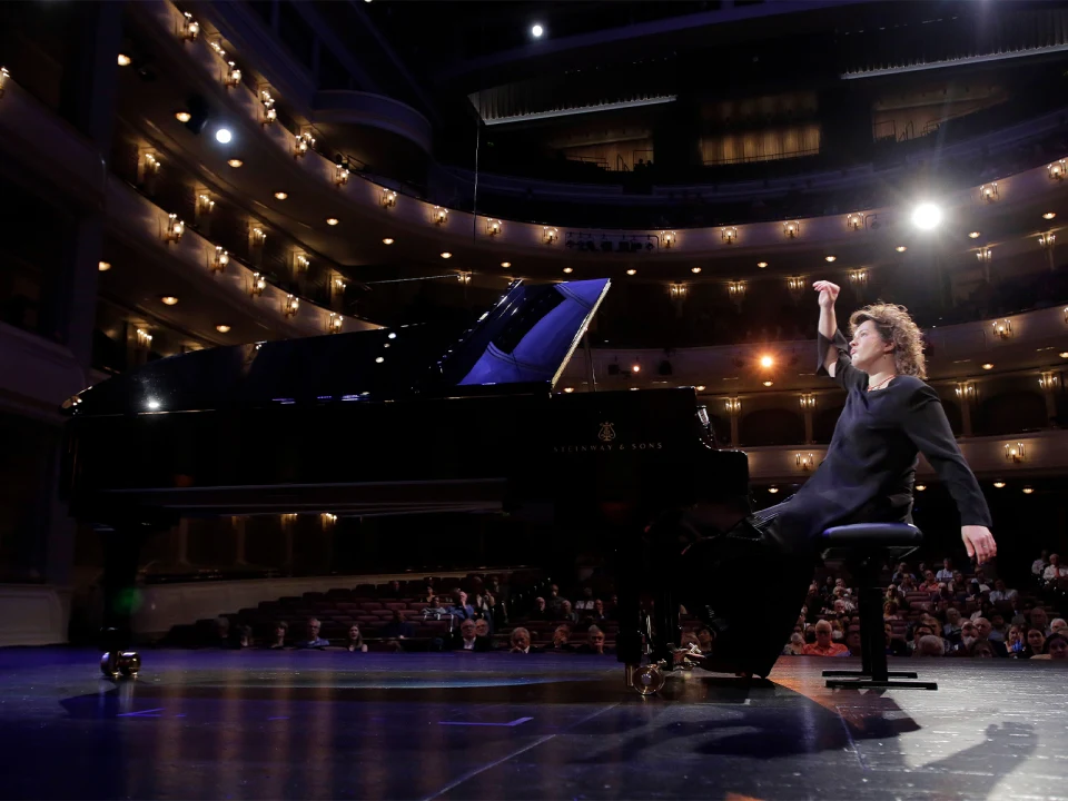 A person wearing a black outfit is performing on a grand piano on a stage in a large, well-lit auditorium with an audience seated in the background.