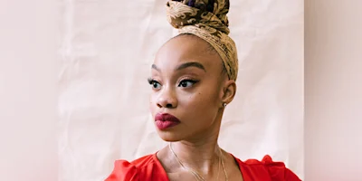 A woman in a red top with a patterned headscarf and bold red lipstick looks to the side against a plain, light-colored background.