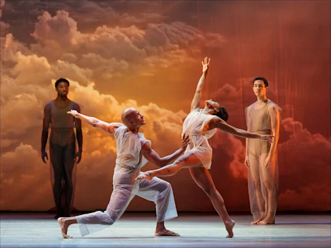 Four dancers perform on stage in sheer costumes against a cloudy sky backdrop; two are in dynamic poses, while two stand in the background observing.