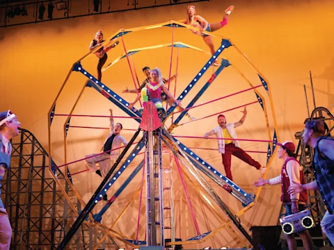 Performers in 1980s-style clothing pose on a colorful Ferris wheel set piece on stage.