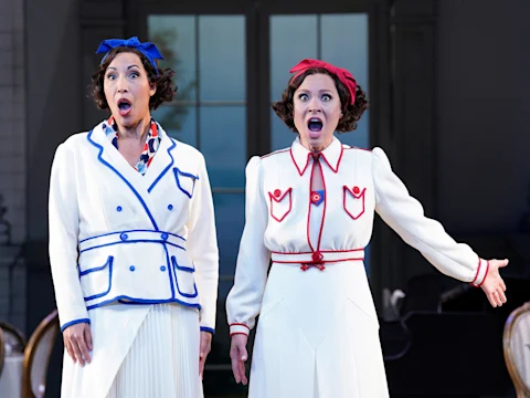 Two women in white outfits with blue and red accents sing on stage, with open mouths and expressive gestures, standing in front of a large window.