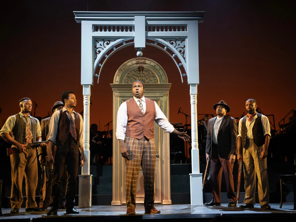 Stage scene with five men in period costumes near an ornate archway. One man stands center, gesturing, while others surround him. The background features a reddish-orange gradient.