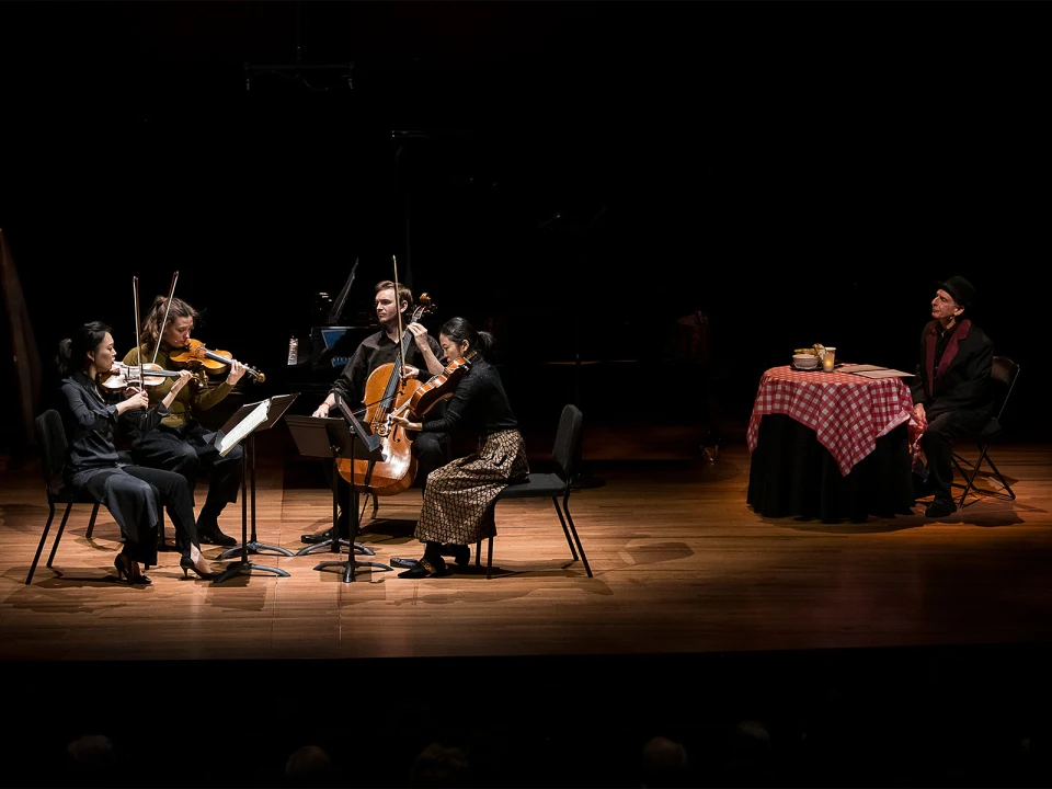 A string quartet performs on a dimly lit stage while a person in a suit and hat sits at a table with a red checkered tablecloth at the side.