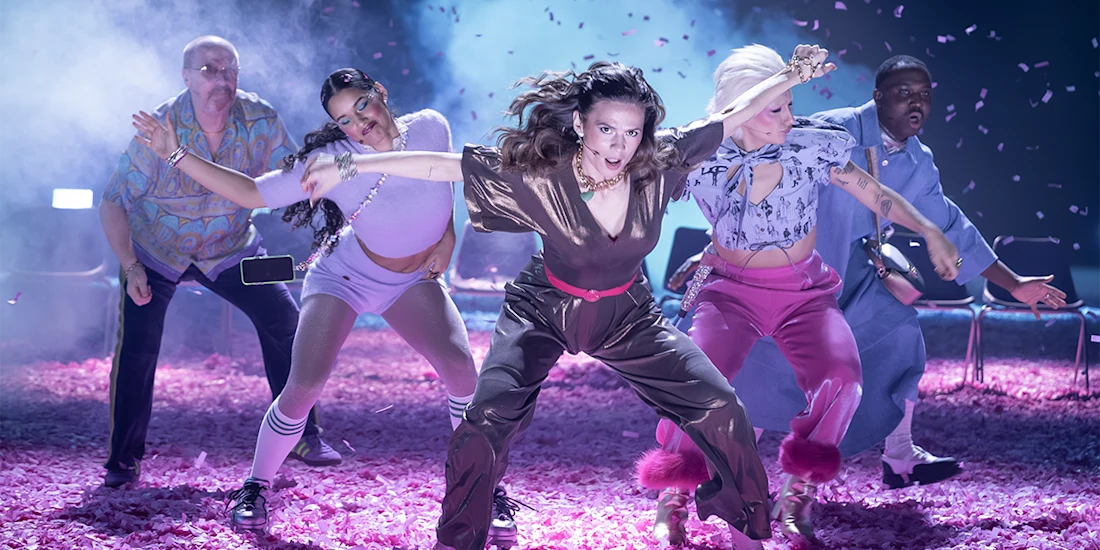 Five people perform a dynamic dance routine on a stage covered in pink confetti, with dramatic lighting and energetic poses.