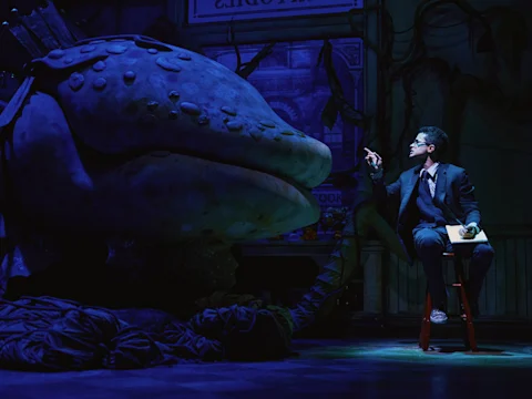 A man in a suit sits on a stool holding papers, facing a large, puppet-like plant with an open mouth on a dimly lit stage.