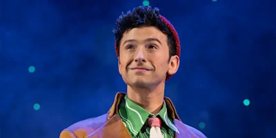 A person wearing a red knit hat, green striped shirt, and brown coat stands in front of a blue background with small lights.