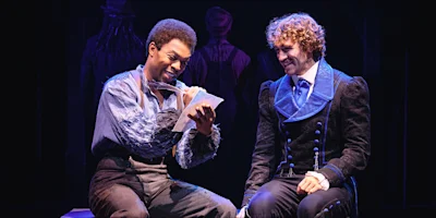 Two actors in period costumes sit on stage; one writes with a quill on paper while the other watches him and smiles.