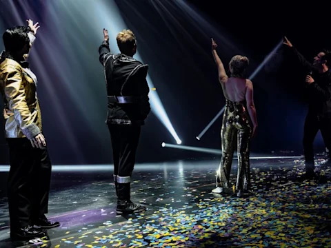 Four performers stand on a confetti-covered stage under spotlights, facing the audience with one arm raised.